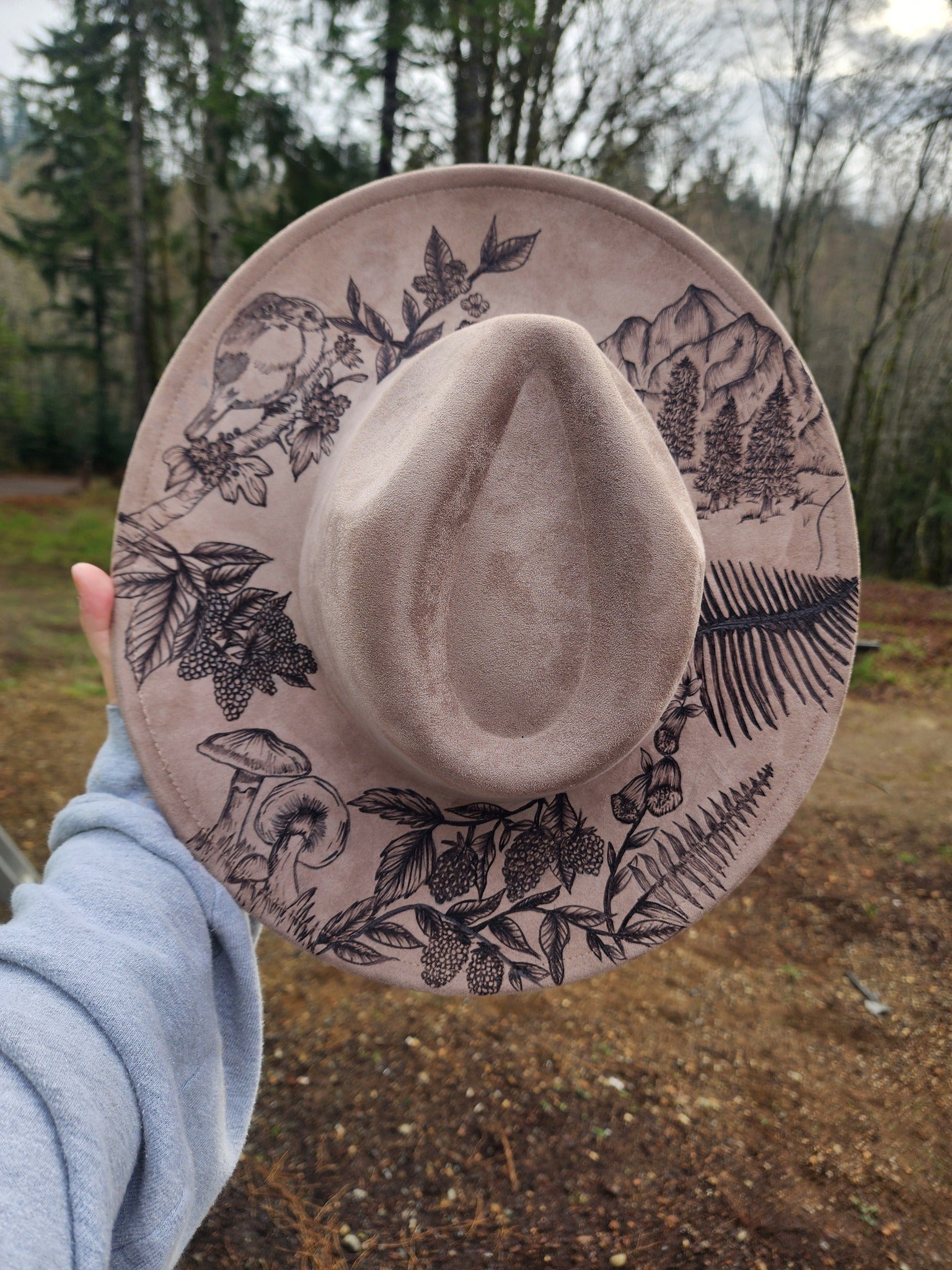 Beige hat with black floral and nature-themed patterns held in front of a forest background
